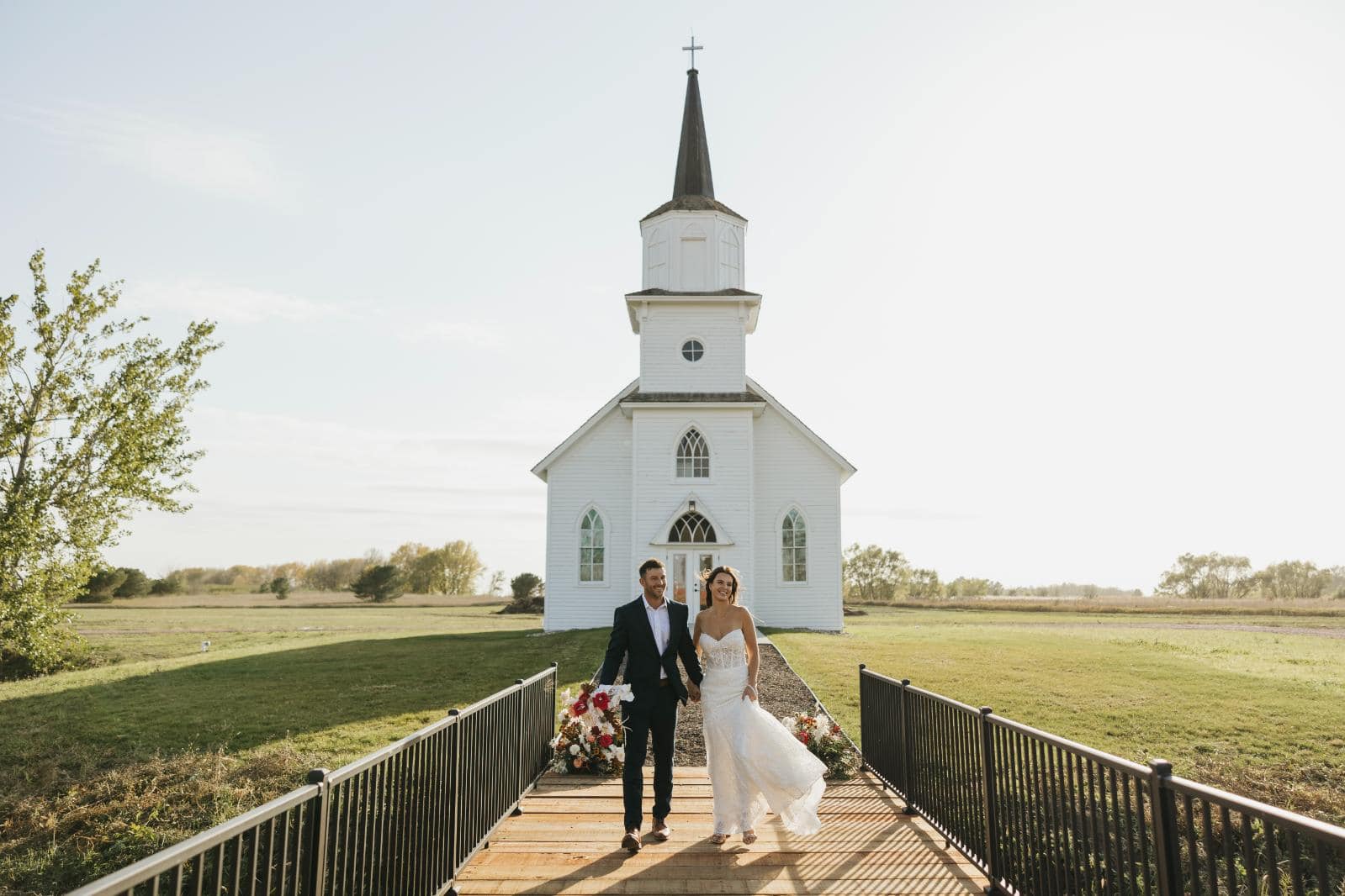In this serene image, a newlywed couple walks hand in hand away from a quaint, traditional white chapel, symbolizing their first steps together in marriage. The chapel, with its classic steeple and arched windows, sits elegantly against a clear blue sky. The surrounding landscape, open and tranquil, suggests a peaceful rural setting. This picturesque moment captures the essence of a joyful and intimate wedding day.